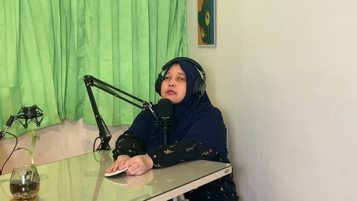 Seorang wanita berhijab biru dan headphone berbicara ke mikrofon di meja, tampak sedang merekam podcast. Di latar belakang ada tirai hijau dan tanaman kecil.